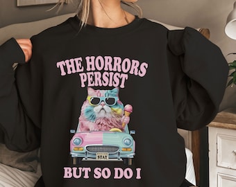 May include: Black sweatshirt with a pink and blue cat wearing sunglasses and holding an ice cream cone. The cat is driving a pink and blue car with the text "The Horrors Persist But So Do I".