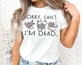 May include: White t-shirt with a graphic of three possums in different poses and the text "Sorry. Can't. I'm Dead."