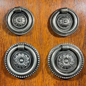 May include: Four antique silver-toned drawer pulls with a floral design. Each pull features a round ring handle and a decorative center with a flower pattern. The pulls are set against a warm, wooden background.
