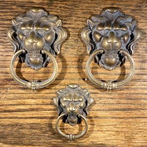 May include: Three antique brass lion head door knobs with ring pulls. The lion heads are detailed with a mane and have a fierce expression. The rings are circular and have a smooth finish.