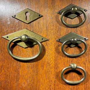 May include: Antique brass hardware set including diamond-shaped backplates and ring pulls. The set includes a keyhole escutcheon. The hardware is displayed on a wooden surface.