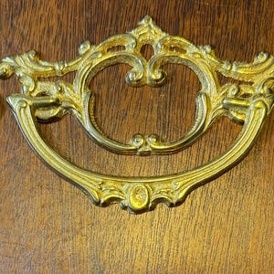 May include: A gold-toned ornate drawer pull with a decorative design. The pull is attached to two screws and comes with two matching decorative screws.