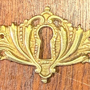 May include: A gold-toned metal keyhole cover with a decorative floral design. The cover has a keyhole in the center and a detailed, ornate design.