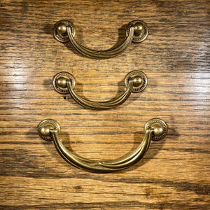 May include: Three gold-toned drawer pulls with a curved design. The pulls are arranged in a vertical row, with the smallest pull at the top and the largest pull at the bottom.