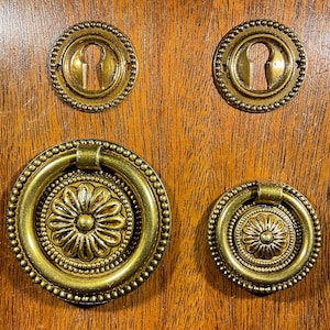 May include: Four antique brass hardware pieces: two keyhole covers and two ring pulls. The keyhole covers are round with a beaded edge. The ring pulls have a floral center design and a ring handle. All pieces are set against a wood grain background.