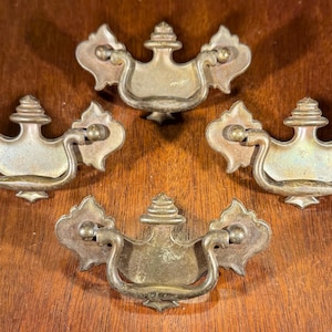 May include: Four antique brass cabinet pulls with ornate designs. Each pull has a central loop and decorative accents.