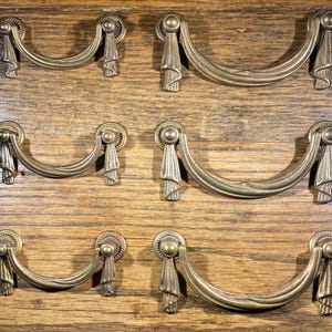 May include: Six antique brass drawer pulls with a decorative ribbon design. The pulls are in various sizes and are mounted on a wooden surface.