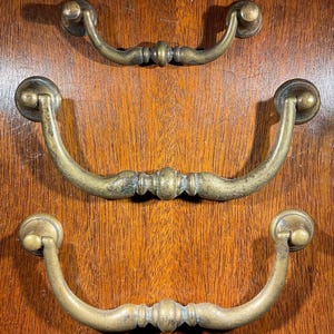 May include: Three antique brass drawer pulls with a curved, arched design. The pulls have a decorative, ornate knob on each end.