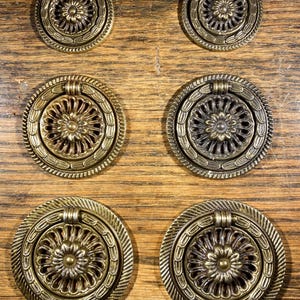 May include: Six antique brass drawer pulls with a floral design. Each pull is circular with a decorative ring around the edge and a small handle at the top. The pulls are mounted on a wooden surface.