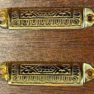 May include: Two gold-colored cabinet pulls with ornate, symmetrical designs. Each pull has a rectangular center with decorative patterns and rounded ends with screw holes. The pulls are displayed on a wooden surface.