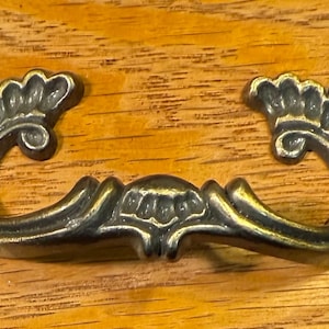 May include: A decorative brass drawer pull with a floral design. The pull has a curved shape and is attached to a wooden surface.