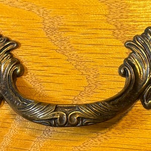 May include: A decorative, ornate, gold-toned metal drawer pull with a leaf design.