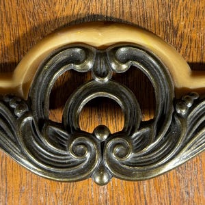 May include: Ornate, antique-style drawer pull with a cream-colored base and dark bronze-colored metal detailing. The design features scrolling flourishes and a central circular element, set against a wooden surface.