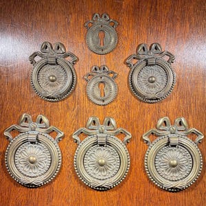 May include: Antique brass drawer pulls and keyhole covers. The drawer pulls have a round design with a floral center and a decorative bow at the top. The keyhole covers are also brass and have a similar design. The hardware is set against a wood background.