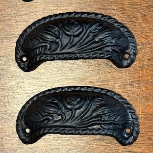 May include: Four black cast iron drawer pulls with a floral design. Each pull has a curved shape with a rope-like border and two holes for mounting. The pulls are arranged vertically on a wooden surface.