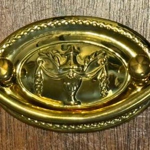 May include: A gold-colored, oval-shaped drawer pull with an ornate design. The center features a decorative urn with draped accents. The pull has rounded ends and a beaded border, adding a touch of elegance to furniture.