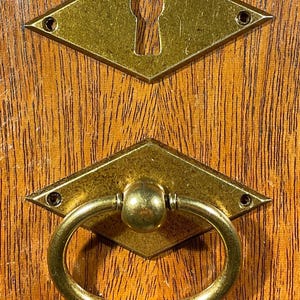 May include: Close-up of two antique brass hardware pieces. The top piece is a diamond-shaped escutcheon with a keyhole. The bottom piece is a diamond-shaped pull with a ring handle. Both pieces are mounted on a wooden surface.