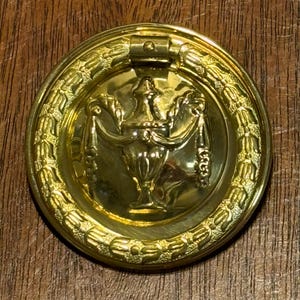 May include: A round, gold-colored cabinet pull with an ornate design. The pull features a central decorative element with a vase shape and draped accents, surrounded by a laurel wreath border. The hardware is likely made of brass or a similar metal.