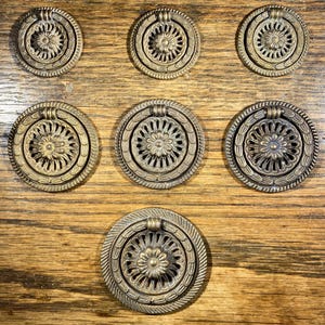 May include: Seven antique brass cabinet knobs with a decorative floral design. The knobs have a ring pull and are arranged in two rows on a wooden surface.