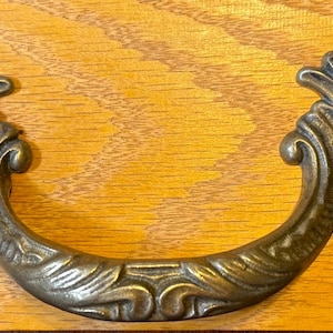 May include: Ornate bronze drawer pull with a curved handle and decorative leaf and scroll design. The hardware is mounted on a light-colored wooden surface, showcasing its intricate details and vintage aesthetic.