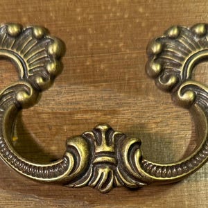 May include: A gold-toned ornate drawer pull with a decorative, swirling design. The pull is made of metal and has a classic, elegant style.
