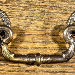 May include: A brass drawer pull with a decorative, ornate design. The pull has a curved shape and two round knobs on each end. The pull comes with two screws and two nuts.