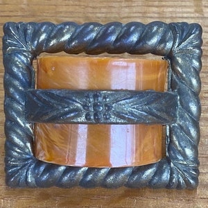 May include: A square, antique-style candle holder with a twisted rope-like frame and a decorative band. The holder contains an orange candle. The metal frame has a dark, aged finish, and the candle is visible through the frame.