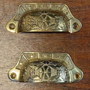 Puede incluir: Dos tiradores de latón antiguos. Cada tirador presenta un diseño floral y geométrico ornamentado. Los tiradores tienen forma semicircular con orificios de montaje en cada extremo. El latón tiene un tono dorado cálido.