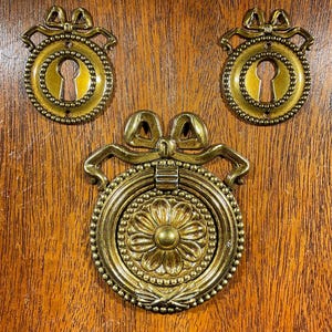 May include: Three antique brass hardware pieces. Two keyhole covers with bow details and a central pull with a floral design. The hardware is set against a warm, wooden background.