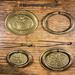 May include: Set of five gold-toned metal drawer pulls with an oval shape and a cherub design. Each pull has a raised edge and a floral pattern around the perimeter.