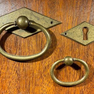 May include: Antique brass hardware set, including a diamond-shaped escutcheon with a keyhole, a large ring pull, and a smaller ring pull. The hardware is mounted on a wooden surface with a visible wood grain.