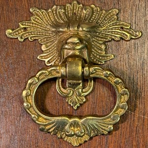 May include: A gold-toned door knocker with a detailed design of a face with a beard and a ring pull. The knocker is attached to a wooden door.