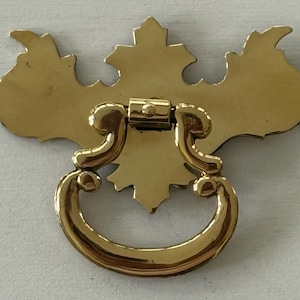 May include: A gold-colored drawer pull with a decorative backplate and a curved handle. The backplate has a stylized leaf design. The handle is a semi-circular shape. The hardware is made of metal.