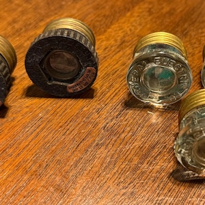May include: Six glass and metal fuse holders. The fuse holders are clear glass with a brass colored metal top. The glass is textured and has a raised ring around the top. The metal top has a raised ring around the bottom. The fuse holders are arranged in a row on a wooden surface.