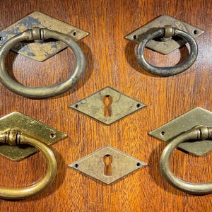 May include: Antique brass drawer pulls and keyhole covers. The pulls have a diamond-shaped backplate and a circular handle. The keyhole covers are diamond-shaped with a keyhole in the center. The hardware is mounted on a wooden surface.