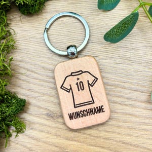 Könnte beinhalten: Holzschlüsselanhänger mit einem silbernen Schlüsselring. Der Schlüsselanhänger zeigt ein lasergraviertes Design eines Fußballtrikots mit der Nummer 10 und dem Text "WUNSCHNAME".