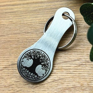 Könnte beinhalten: Silberfarbener Schlüsselanhänger mit einem eingravierten Lebensbaum-Design auf der Vorderseite. Der Schlüsselanhänger hat einen geteilten Ring zum Anbringen von Schlüsseln.