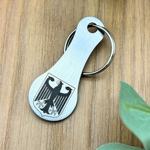 Könnte beinhalten: Ein silberfarbener Metall-Schlüsselanhänger mit einem deutschen Wappen-Emblem. Das Emblem zeigt einen schwarzen Adler auf einem Schild. Der Schlüsselanhänger hat einen Schlüsselring, der an einem tropfenförmigen Anhänger befestigt ist. Der Hintergrund ist eine Holzoberfläche.