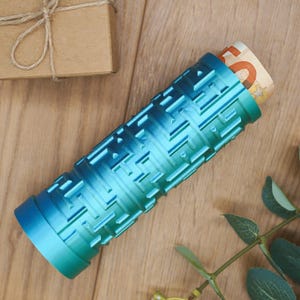 Könnte beinhalten: Ein zylindrischer, blau-grüner Geldhalter mit geometrischem Muster. Der Halter enthält eine aufgerollte Banknote. Im Hintergrund befindet sich eine kleine, verpackte Geschenkbox.
