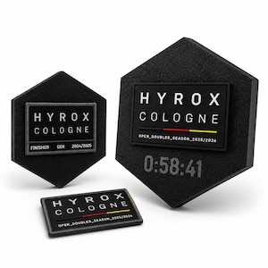 Könnte beinhalten: Drei schwarze sechseckige HYROX Köln Finisher-Auszeichnungen. Die größte zeigt die Zeit "0:58:41". Jede verfügt über ein rechteckiges Patch mit dem HYROX Köln Logo, Eventdetails und dem Text "OPEN_DOUBLES_SEASON_2025/2026".