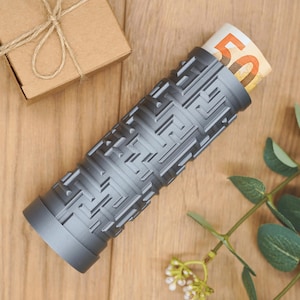Könnte beinhalten: Ein grauer zylindrischer Behälter mit einem Labyrinthmuster, der einen gefalteten Fünfzig-Euro-Schein enthält. Eine kleine braune Geschenkbox mit Bindfaden befindet sich im Hintergrund. Der Behälter steht auf einer Holzoberfläche.