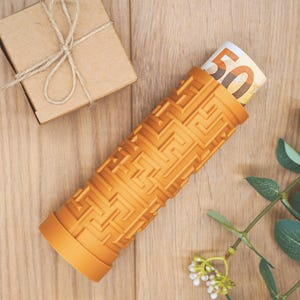Könnte beinhalten: Ein orangefarbener zylindrischer Behälter mit einem Labyrinthmuster, der einen aufgerollten 50-Euro-Schein enthält. Eine kleine braune Geschenkbox, die mit Bindfaden gebunden ist, befindet sich im Hintergrund. Der Behälter steht auf einer Holzoberfläche.