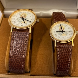 May include: Two gold-tone wristwatches with brown leather bands in a brown velvet box. The watch faces feature a white background with gold Roman numeral hour markers and a depiction of Jesus Christ.