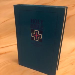 Könnte beinhalten: Eine tealblaue Hardcover-Bibel mit einem weißen Kreuz und den Worten "HOLY BIBLE" auf dem Einband.