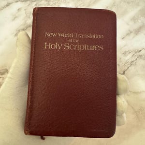 May include: A burgundy-colored book with the title "New World Translation of the Holy Scriptures" in gold lettering. The book is small and rectangular, with a textured cover. The image is a close-up shot.