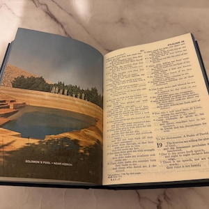 May include: An open book with a blue cover, showing text and an illustration of Solomon's Pool near Hebron. The illustration depicts a pool of water in a landscape with trees and a blue sky. The text is in English.
