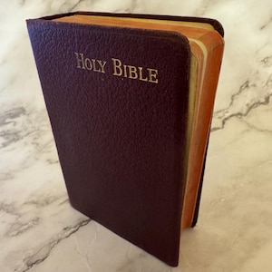 Può includere: Una Sacra Bibbia rilegata in pelle bordeaux con scritte dorate. Le pagine del libro hanno una tonalità dorata e la copertina ha un aspetto strutturato. Il libro è leggermente aperto, rivelando le pagine invecchiate.