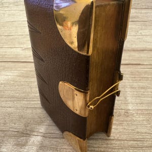 Puede incluir: Una caja rectangular diseñada para parecer un libro antiguo con una cubierta texturizada de color marrón oscuro. La caja cuenta con protectores de esquina de metal dorado pulido y un mecanismo de cierre de metal dorado en el lateral.