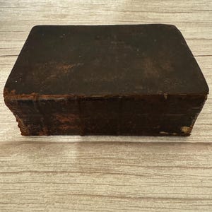 Peut inclure: Un livre ancien rectangulaire doté d'une couverture en cuir marron foncé patiné. Le dos présente des nerfs saillants et montre des signes d'usure importants, notamment des bords effilochés et des surfaces éraflées, témoignant de son âge.