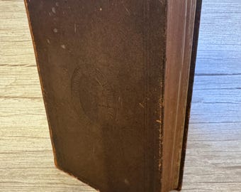 May include: A vintage, rectangular, dark brown hardcover book with a worn, textured surface. The front cover features a central circular embossed design with faint, illegible lettering. The edges of the book show signs of age and wear.